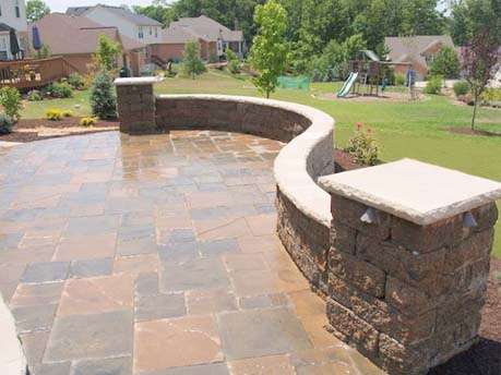 A stone patio with a curved stone wall, crafted by expert patio builders, overlooks a grassy backyard with trees, shrubs, and a children’s playground in the distance.