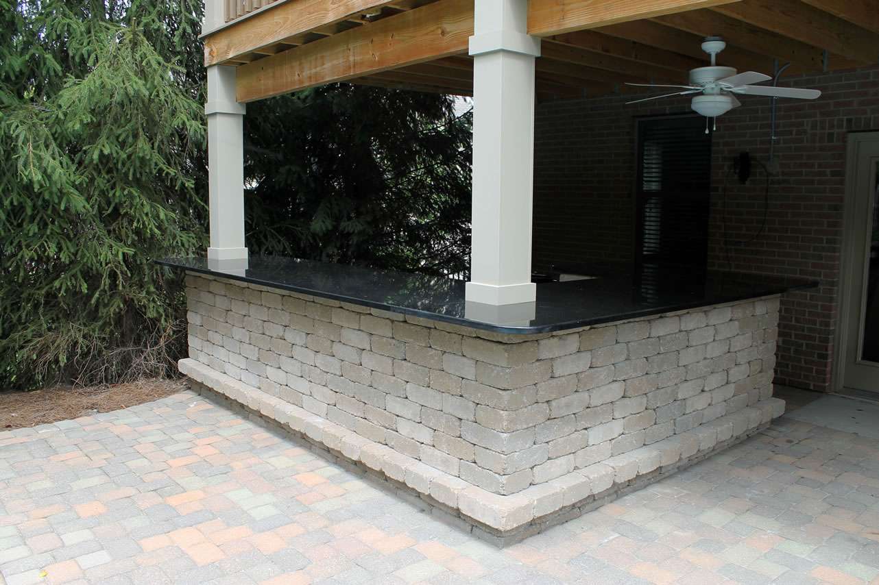 Outdoor bar with a stone base and black countertop under a wooden deck, featuring a ceiling fan and surrounded by brick and paver flooring—expertly crafted by leading patio builders and masonry contractors for stylish outdoor living.