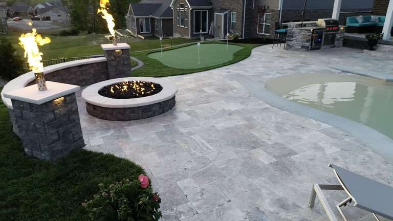 A backyard patio by expert patio builders features a fire pit with flames, a small putting green, and a partially visible pool surrounded by stone and concrete surfaces.