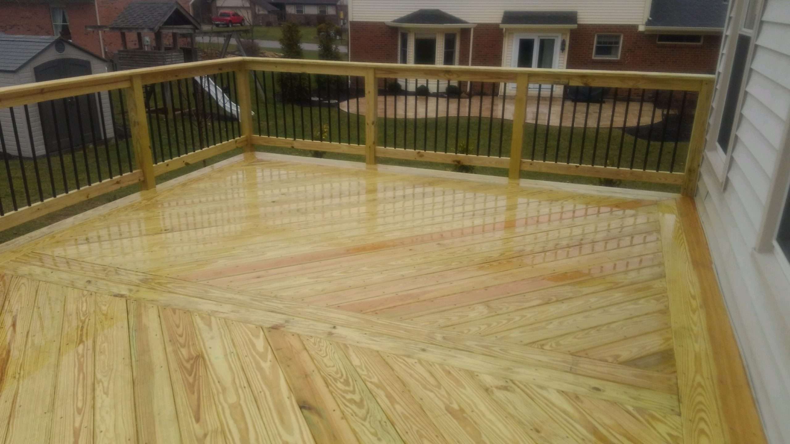 A newly constructed wooden deck with black metal balusters, wet from rain, overlooks a grassy backyard and neighboring houses—perfect for enjoying your outdoor space and ideal inspiration for deck builders or landscaping companies.