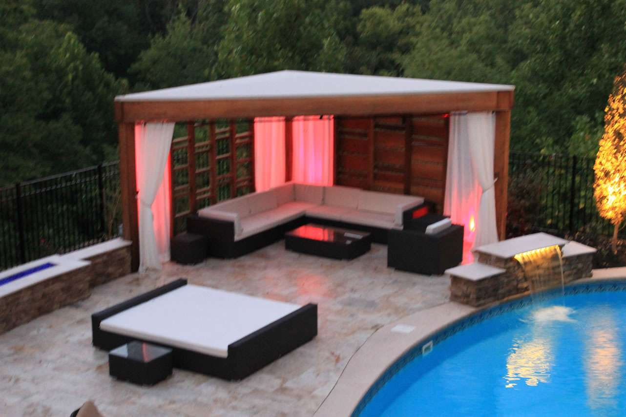 A poolside patio features a covered lounge area with cushioned seating, designed by expert patio builders, with tables and soft lighting beside a curved swimming pool surrounded by trees.
