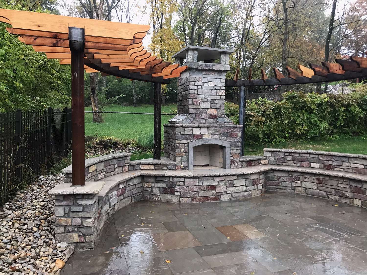 Outdoor stone fireplace with a curved stone bench and wooden pergola, set on a stone patio surrounded by greenery—expertly crafted by patio builders and landscaping companies for a perfect outdoor retreat.