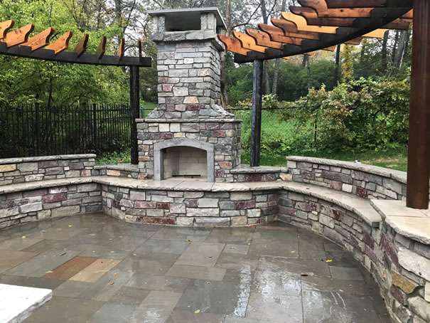 Stone outdoor fireplace with curved stone benches, pergolas overhead by expert pergola builders, and a wet stone patio, set in a green, fenced backyard. Ideal for gatherings designed by leading patio builders.
