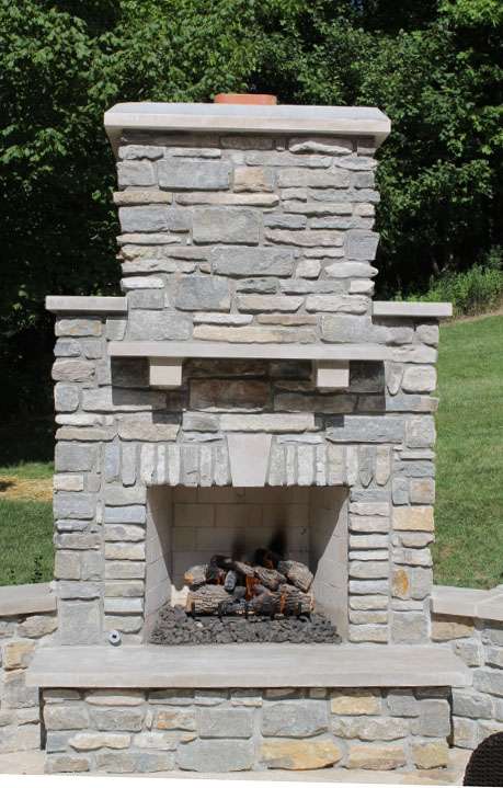 Outdoor stone fireplace with a rectangular chimney, mantel, and artificial logs set inside—perfectly crafted by expert patio builders—and positioned on a patio with grass and trees in the background.