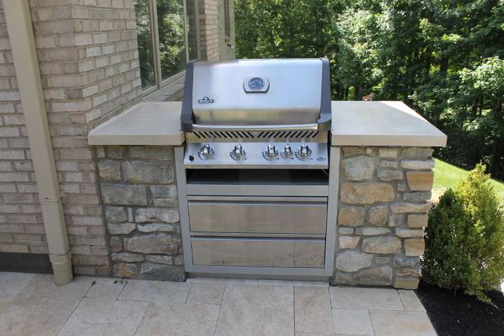 A built-in stainless steel outdoor gas grill with four burners is set into a stone and concrete countertop by expert patio builders on a stylish patio next to a brick house.