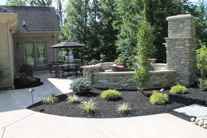 A landscaped backyard patio features stone walls, outdoor seating, a dining table with an umbrella, lush plants and shrubs—expertly designed by professional patio builders and retaining wall contractors.