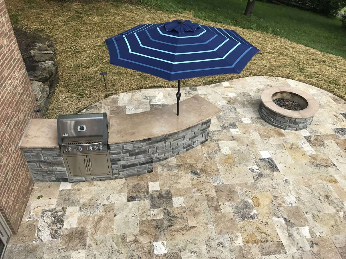 Stone patio with a built-in grill, curved stone counter, blue outdoor umbrella, and a circular fire pit—expertly crafted by top patio builders and masonry contractors for the perfect backyard retreat.