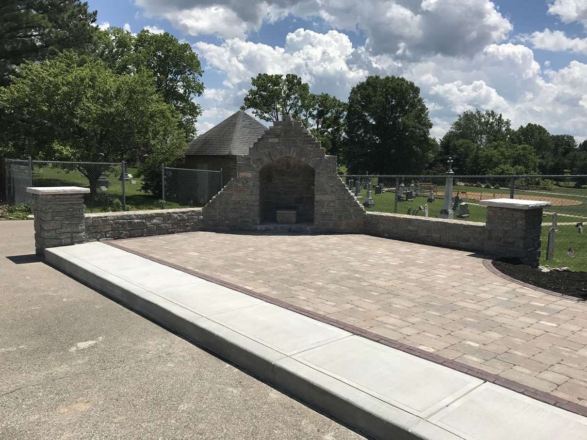 A small stone memorial wall with a central arched alcove stands on a paved area near a cemetery, built by skilled masonry contractors, with trees and a fence in the background.