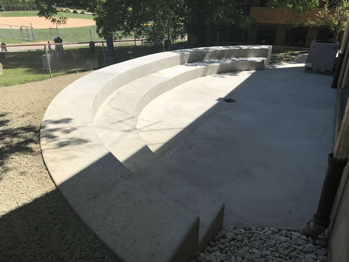 Three curved concrete benches arranged in a semicircle on a paved outdoor area near a building, with landscape stone accents and a sports field visible in the background, ideal inspiration for landscaping companies and patio builders.