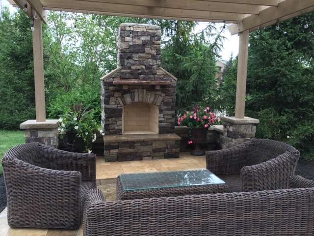 Wicker patio furniture arranged around a glass-top table faces a stone outdoor fireplace under a wooden pergola, crafted by expert patio builders, with lush greenery in the background.