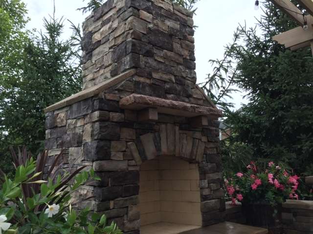 A large outdoor stone fireplace with a tall chimney stands on a patio, surrounded by green trees and flowering plants—an inviting oasis created by expert landscaping companies and patio builders.