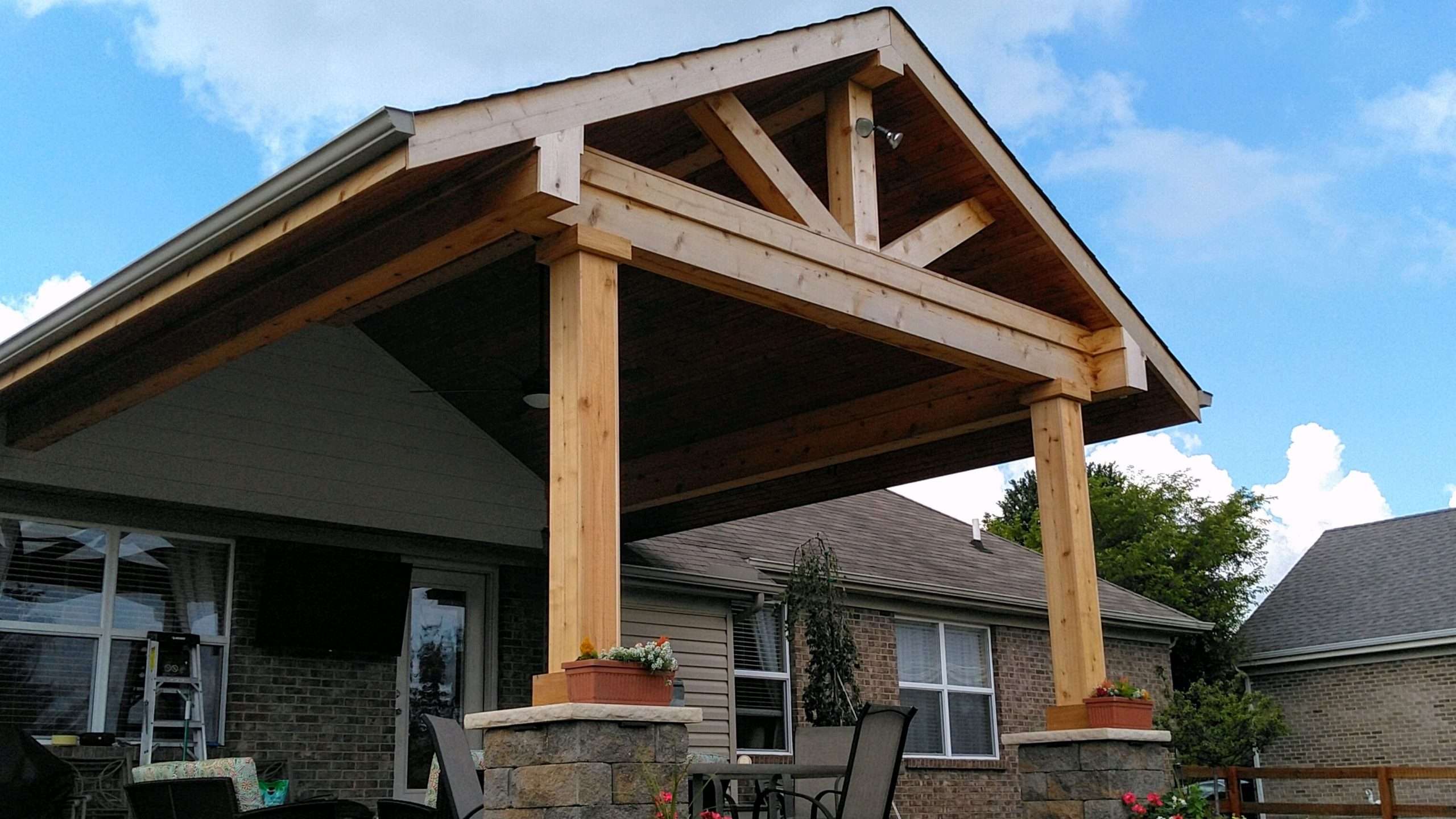 A covered patio with a wooden gable roof supported by stone and wood columns, outdoor furniture, and potted plants, crafted by expert patio builders and masonry contractors, attached to a charming brick house.