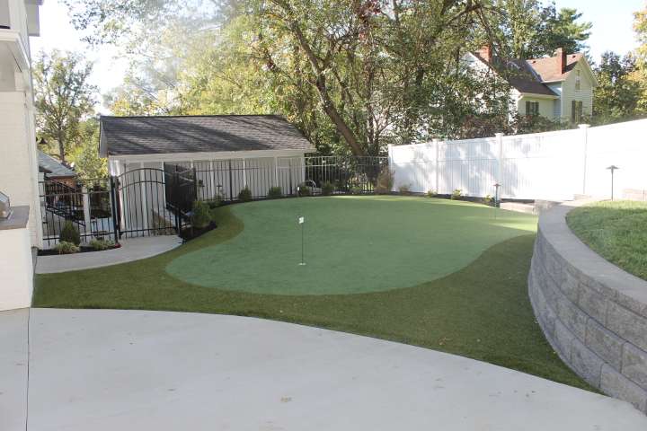 A small backyard putting green with a single flag, surrounded by a white fence, stone retaining wall built by expert retaining wall contractors, trees, and a shed.