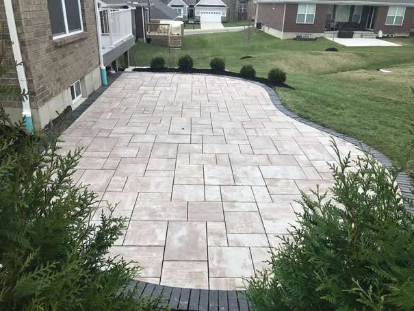 A backyard patio with light-colored rectangular pavers bordered by dark bricks, adjacent to a lawn and surrounded by bushes—an inviting retreat crafted by expert patio builders and landscaping companies.