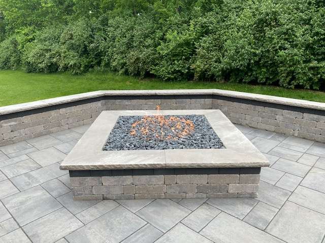 A square stone fire pit with black rocks and a gas flame sits on a paved patio, expertly crafted by outdoor patio builders and surrounded by a low stone wall and lush green trees in the background.