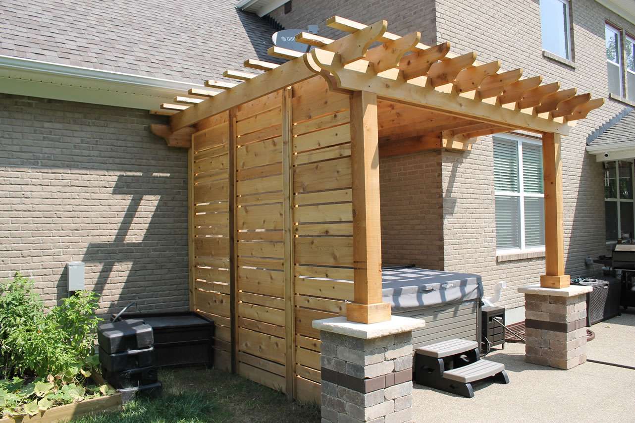 A wooden pergola with privacy panels is attached to a brick house, providing shade over a hot tub on a concrete patio. Gardening containers are visible nearby—perfect inspiration for patio upgrades by expert patio builders or landscaping companies.