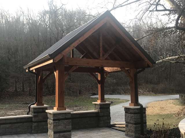 Wooden pavilion with a metal roof and stone pillars stands on a paved area near a gravel driveway and wooded background, showcasing the craftsmanship of expert patio builders and landscape stone specialists.
