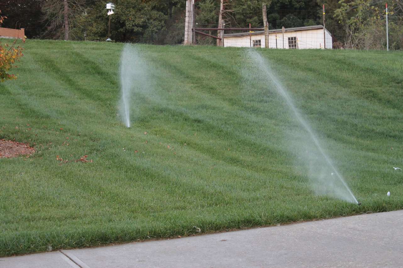 Two lawn sprinklers spray water onto a sloped grassy yard near a concrete walkway, with a shed and trees in the background—perfect inspiration for irrigation system companies or sprinkler system installation experts.