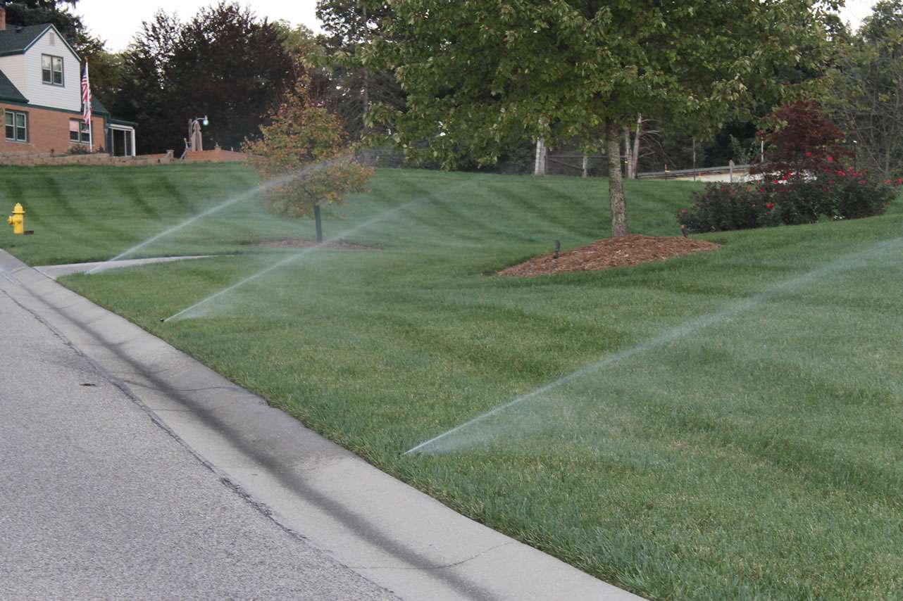 Several sprinklers are watering the grass along the edge of a suburban lawn near a street curb on a cloudy day, showcasing the work of professional irrigation system companies and expert sprinkler system installation.