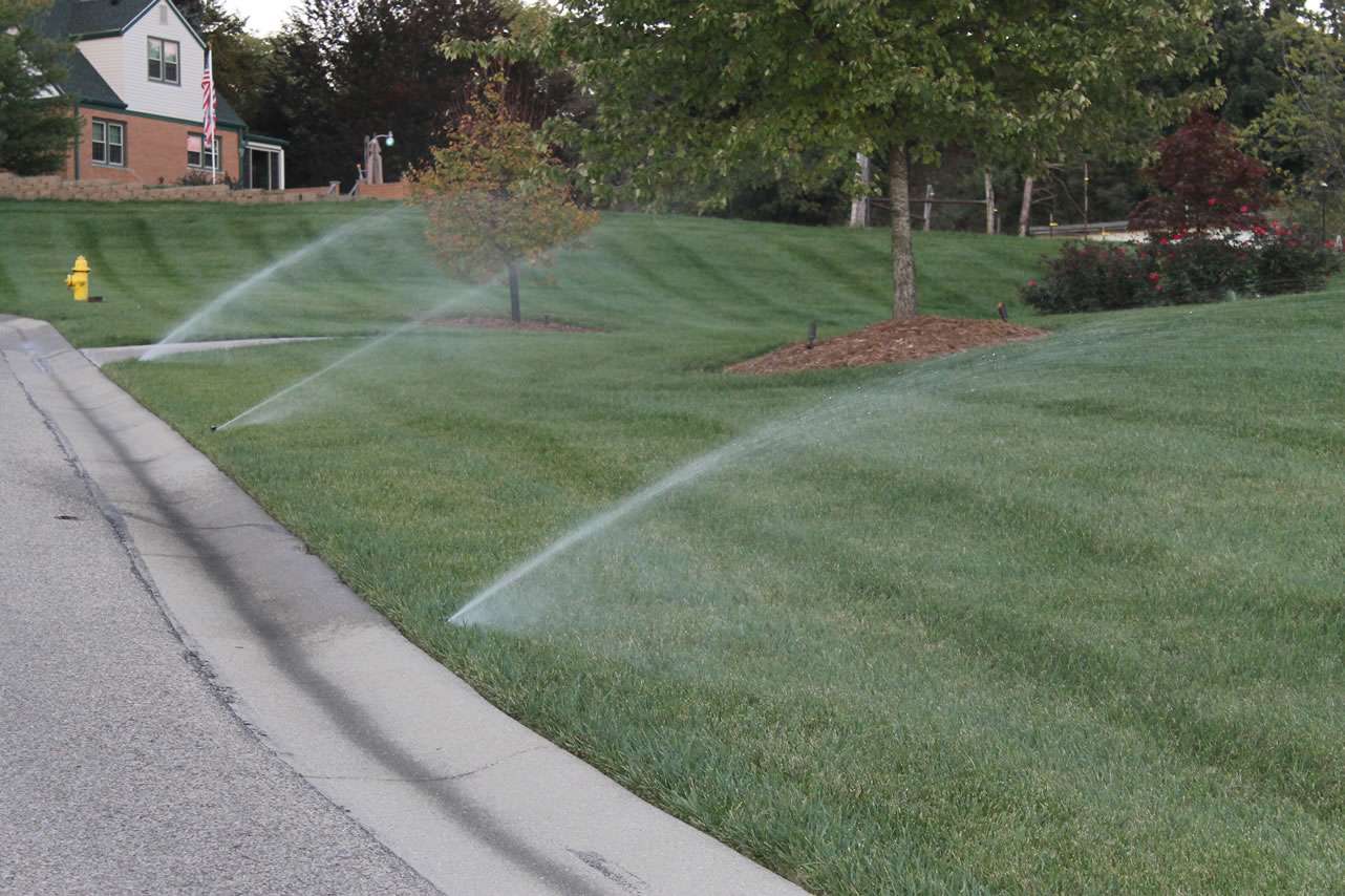 Lawn sprinklers water grass along the edge of a residential street, where houses, trees, and a fire hydrant are visible—perfectly illustrating the work of irrigation system companies and expert sprinkler system installation.