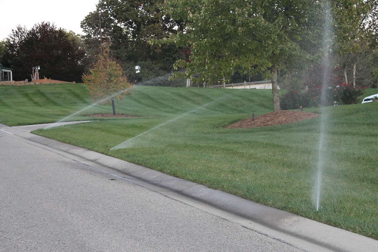 Automatic sprinklers watering a well-maintained lawn beside a street, with trees and landscaped areas visible in the background, showcase the work of top irrigation system companies and expert landscaping companies.