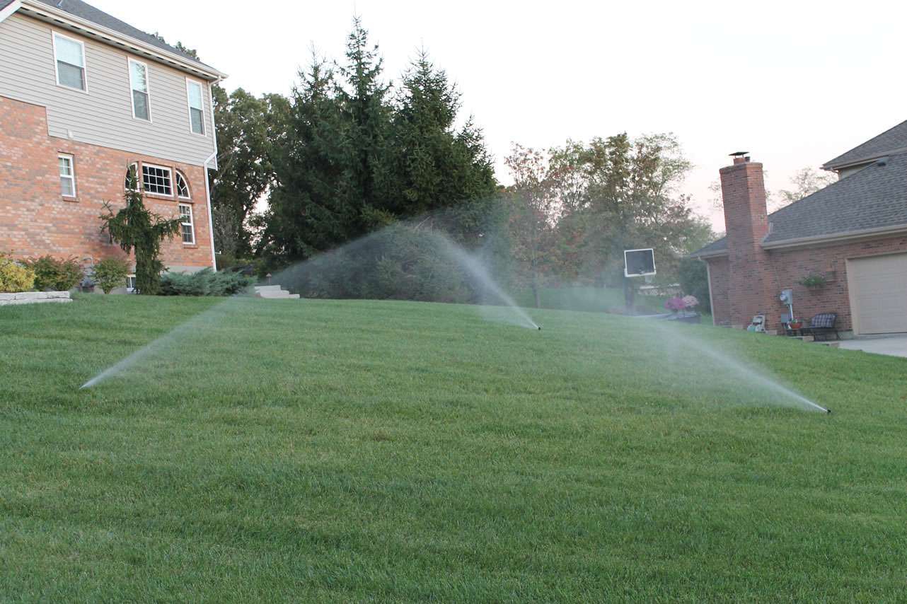 Two sprinklers water a green lawn in front of suburban houses, with trees and a basketball hoop visible in the background—perfect for showcasing the work of local irrigation system companies and sprinkler system installation experts.