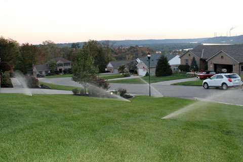 Sprinklers watering the grass on a suburban lawn at dusk, with houses, parked cars, and a hilly landscape in the background—an inviting scene made possible by top irrigation system companies and professional sprinkler system installation.
