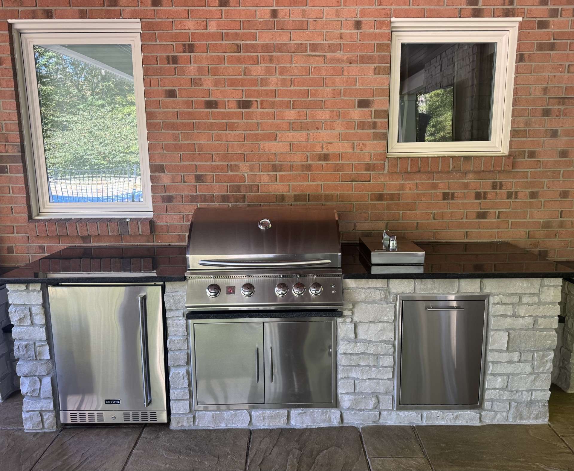 Outdoor kitchen with stainless steel appliances, including a mini fridge, grill, sink, and storage, set in a stone and brick structure under two windows.