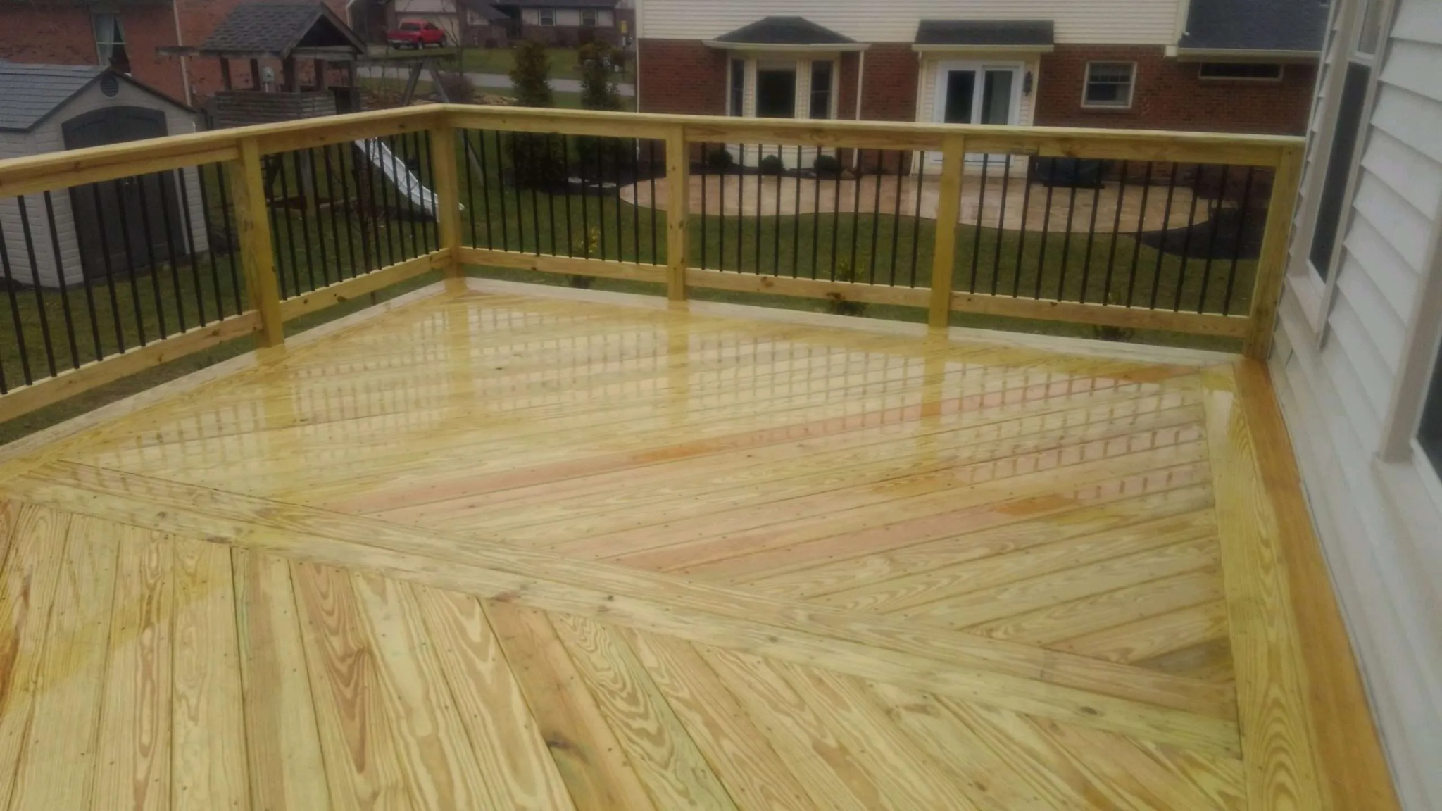Wet wooden backyard deck with black metal railings, overlooking neighboring homes and lawns on an overcast day.