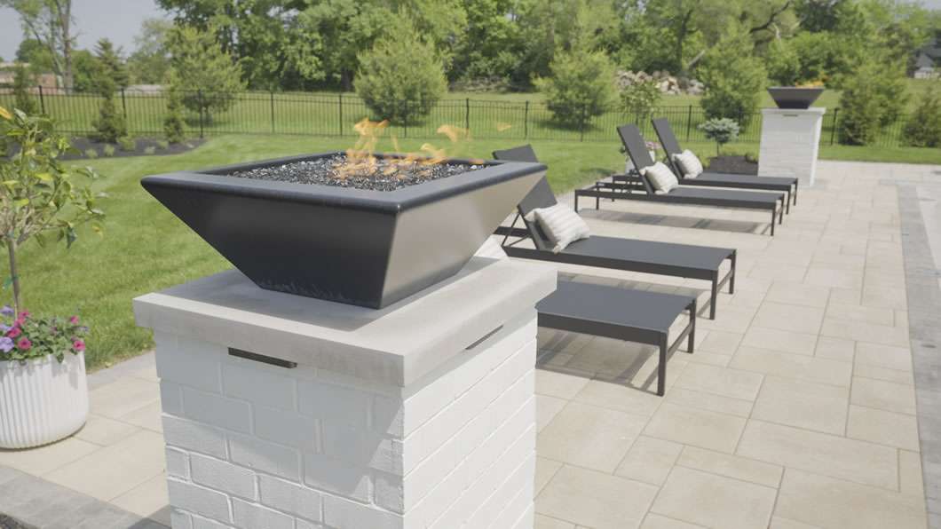 Modern outdoor patio with a black fire bowl on a white brick pedestal and several black lounge chairs arranged in a row beside a landscaped garden.