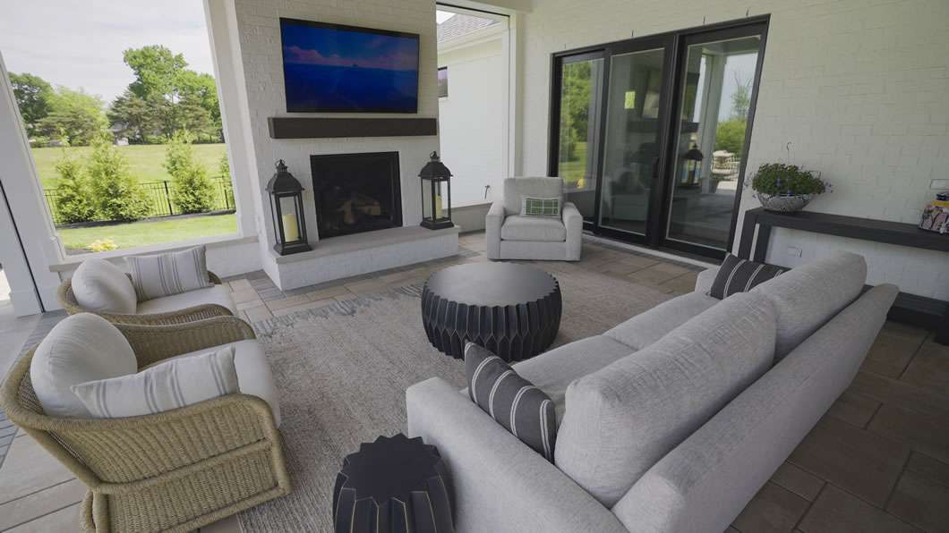 Outdoor patio seating area with a sofa, armchairs, round tables, a rug, fireplace, mounted TV, and large sliding glass doors opening to a grassy yard.