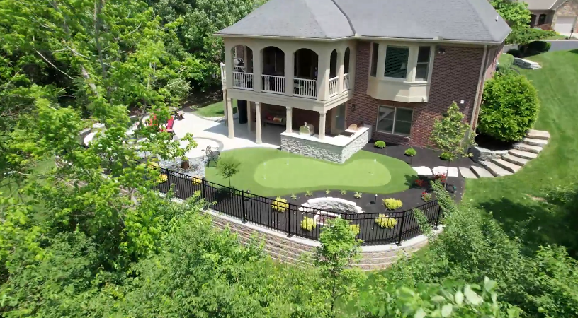 Aerial view of a two-story brick house with a covered patio, landscaped yard, and a small putting green surrounded by a black metal fence and lush greenery. Aerial view of a two-story brick house with a covered patio, landscaped yard, and a small putting green surrounded by a black metal fence and lush greenery.